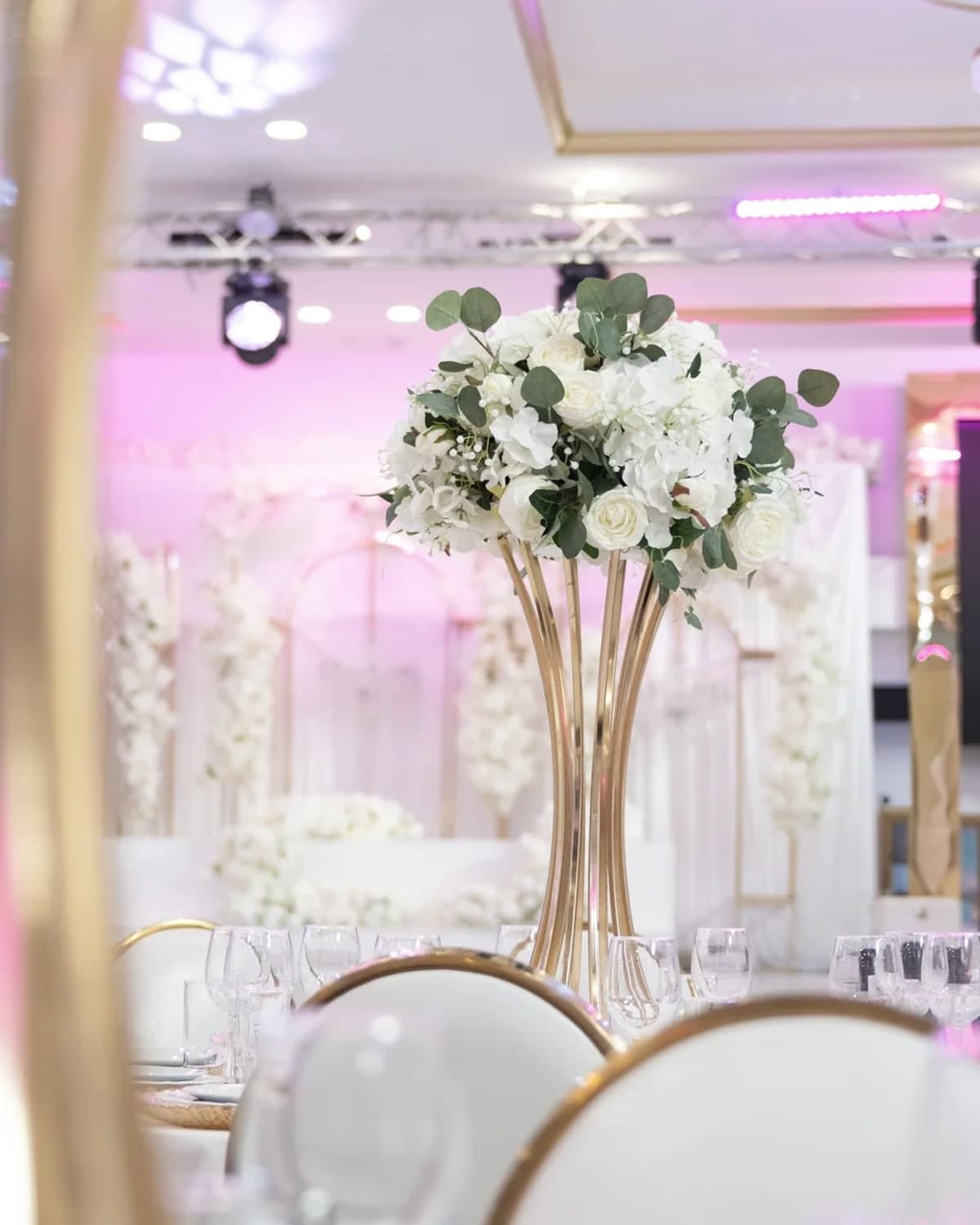 Goldener Blumenständer mit weißen Rosen auf einer gedeckten Tafel, davor Gläser und ein goldener Stuhl